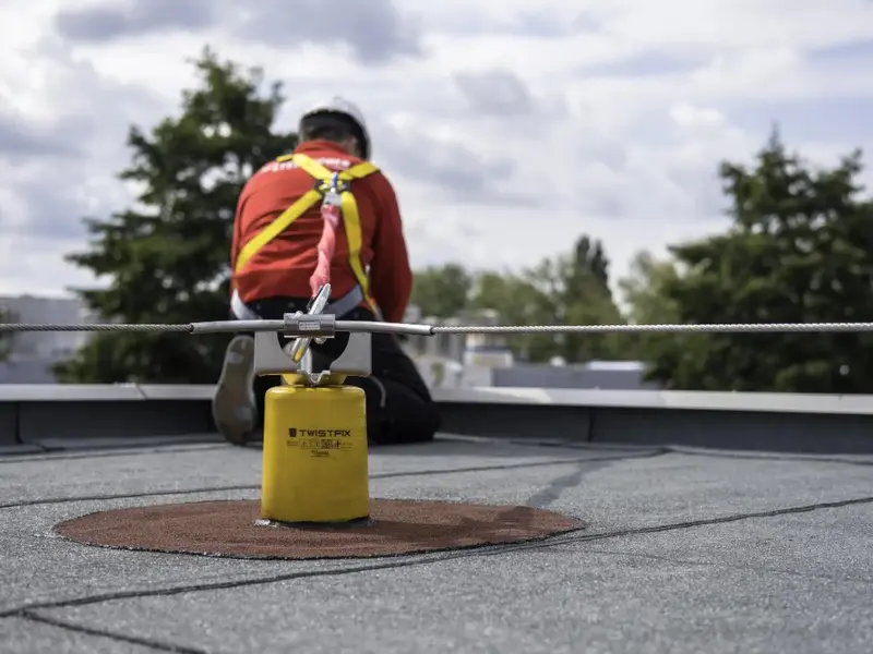 Werker met helm en harnas, aangelijnd aan een staalkabel met een geel ankerpunt op een plat dak