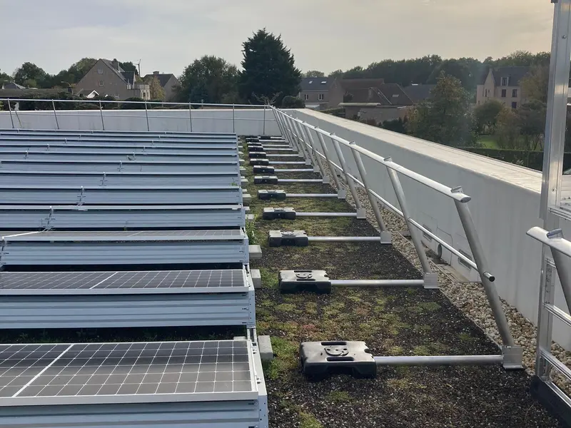 Vrijstaande aluminium leuning met zwarte gewichten op een plat groendak, naast een rij zonnepanelen