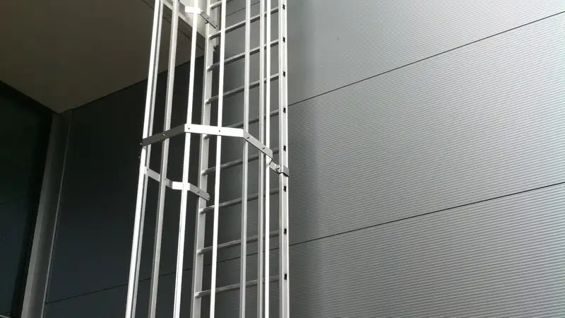 Aluminium kooiladder met een afgesloten inklimbeveiliging onderaan, tegen een grijze gevel van metalen panelen
