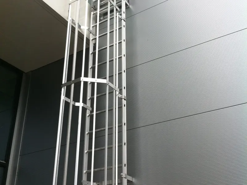 Aluminium kooiladder met een afgesloten inklimbeveiliging onderaan, tegen een grijze gevel van metalen panelen