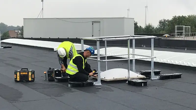 Twee vakmensen plaatsen een hekwerk rond een lichtkoepel op een plat dak.