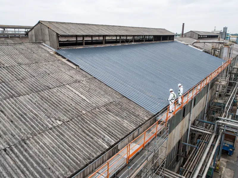 Oud dak van golfplaten naast nieuwe metalen dakplaten op een fabriekshal, met twee werkers in beschermpak op een loopbrug