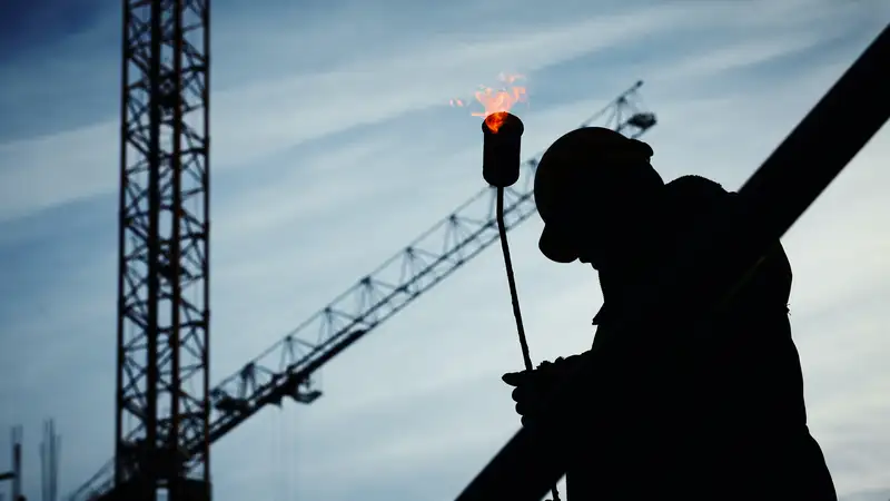 Silhouet van een dakwerker met een brander, met bouwkranen op de achtergrond