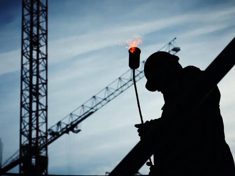 Silhouet van een dakwerker met een brander, met bouwkranen op de achtergrond