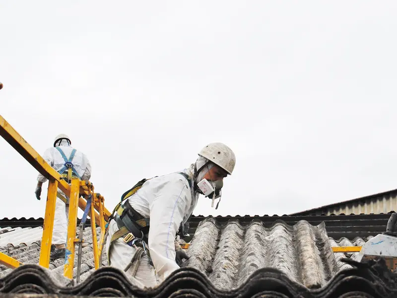 Werkers in beschermpak en harnas halen golfplaten van asbestcement van een dak