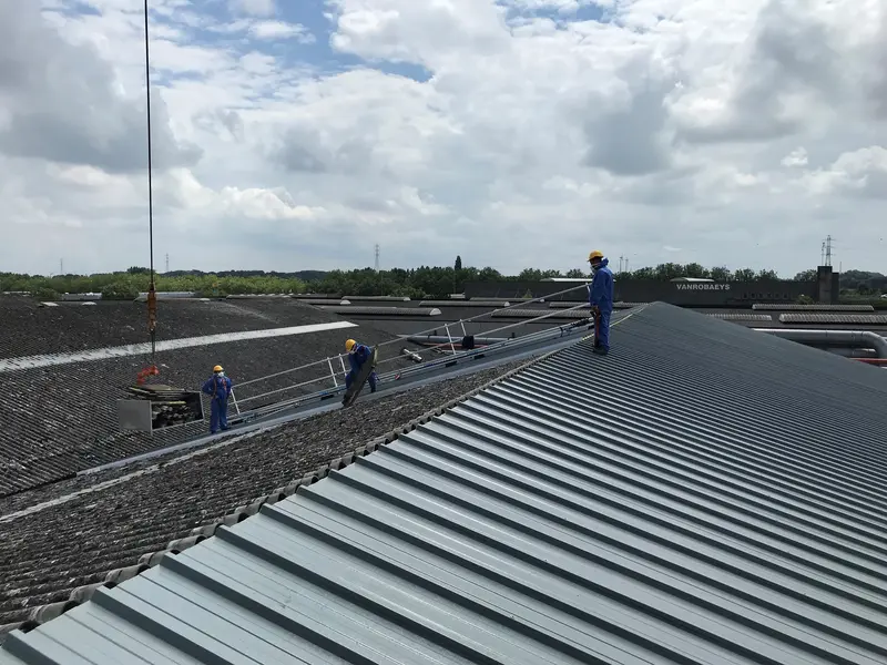 Werkers met helm op een loopbrug met leuning, tussen oude golfplaten en nieuwe metalen dakplaten