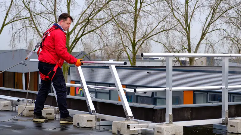 Werker in harnas, aangelijnd, plaatst aluminium hekwerk met betonnen voeten op een plat dak