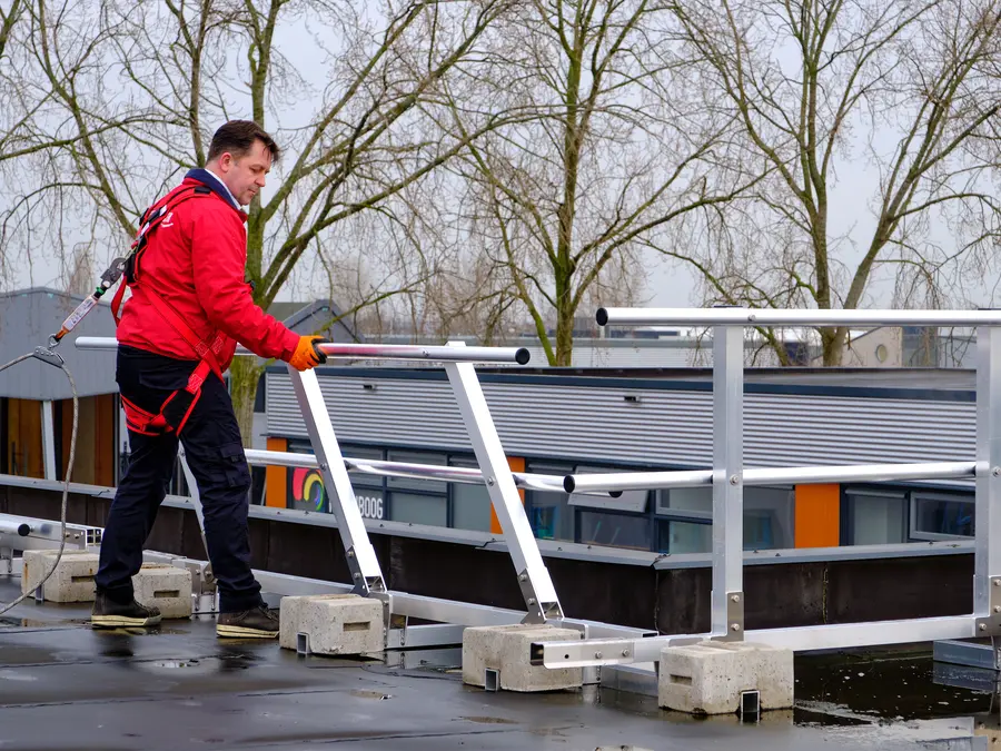 Werker in harnas, aangelijnd, plaatst aluminium hekwerk met betonnen voeten op een plat dak