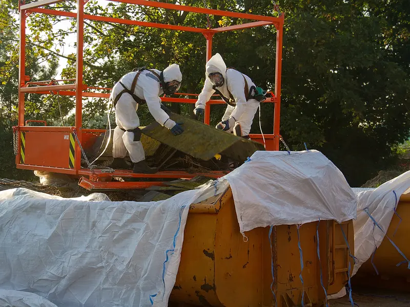 Twee werkers in beschermpak en met masker halen asbestplaten weg vanuit een werkbak