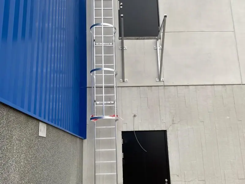 Aluminium kooiladder met kooi tegen een betonnen gevel naast een blauwe hal, met een deur halverwege de gevel.
