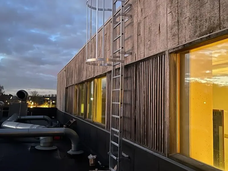 Aluminium kooiladder tegen een betonnen gevel, van een lager plat dak naar het hogere dak, met gereedschap op het dak